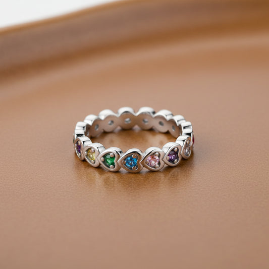 A silver heart-link eternity ring with colorful gemstones, resting on a smooth brown surface.