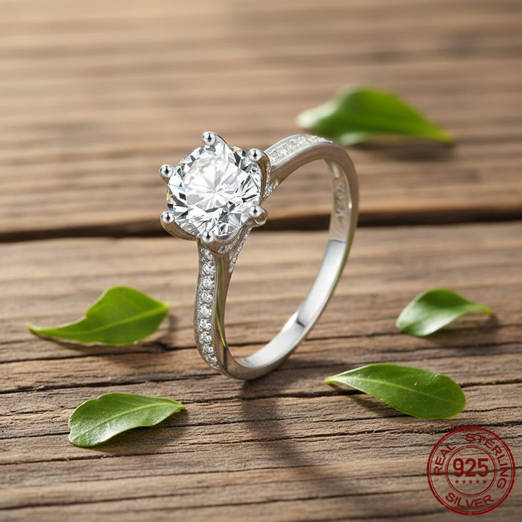 A silver solitaire engagement ring with a round-cut clear gemstone and pavé-set accent stones on the band, displayed on a rustic wooden surface with scattered green leaves. A '925 Sterling Silver' stamp is visible in the bottom right corner.