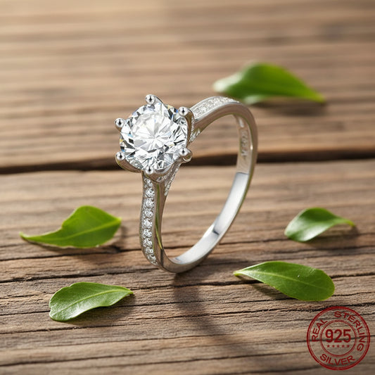 A silver solitaire engagement ring with a round-cut clear gemstone and pavé-set accent stones on the band, displayed on a rustic wooden surface with scattered green leaves. A '925 Sterling Silver' stamp is visible in the bottom right corner.