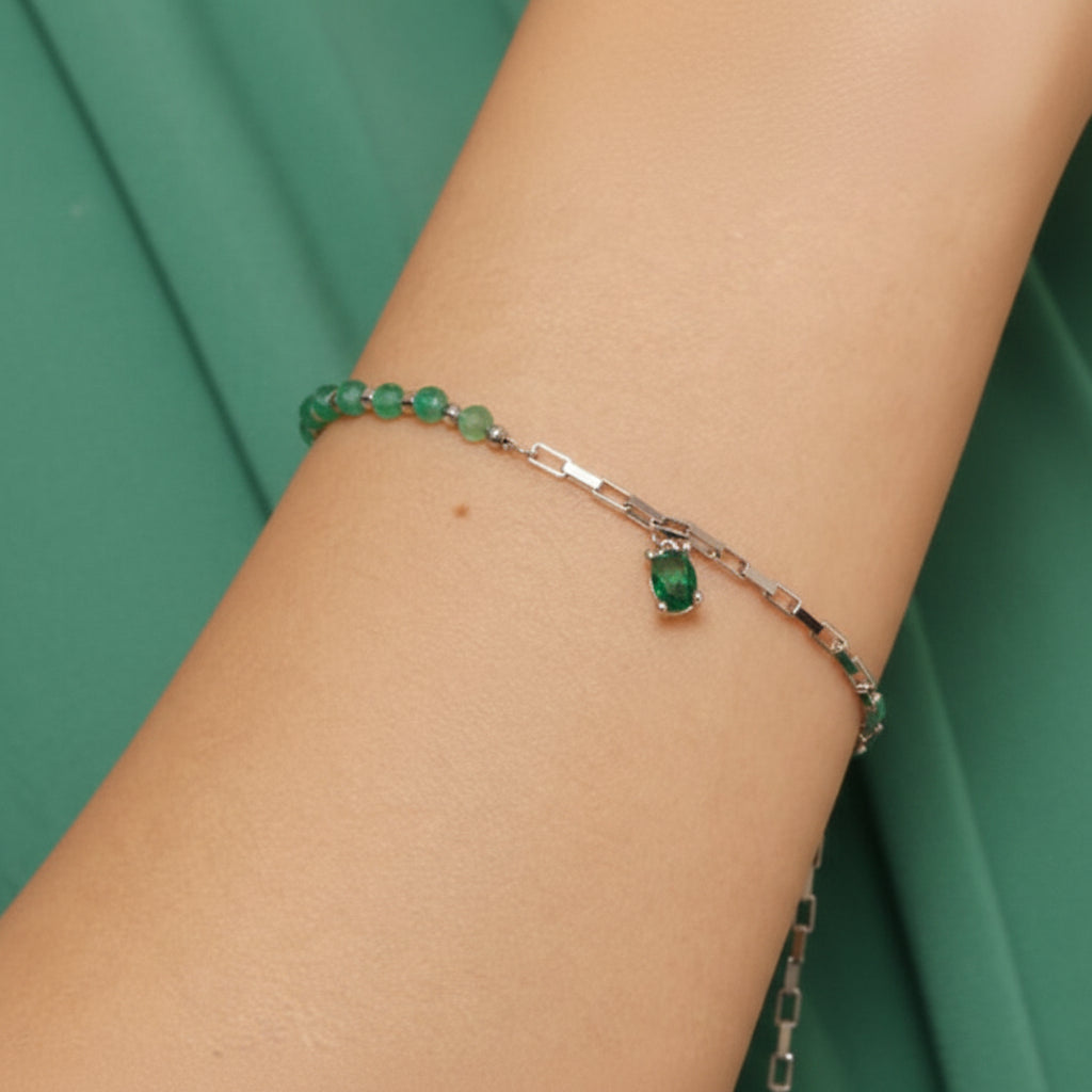 A silver chain bracelet with a dangling emerald-cut green gemstone, worn on a person's wrist against a green fabric background.